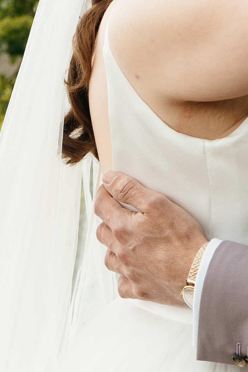 Groom's hand with gold watch resting on bride's white wedding dress and veil during ceremony.