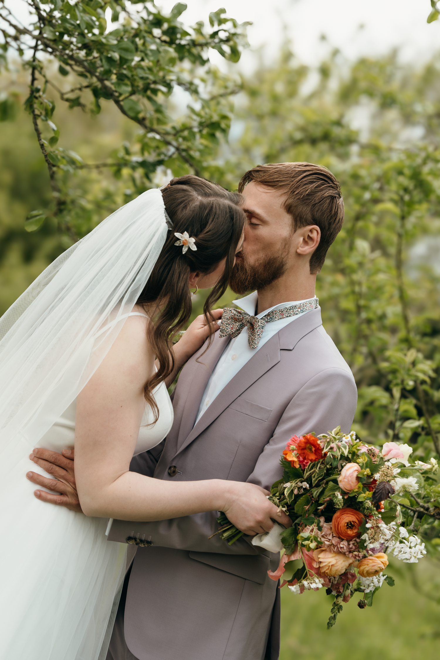 A bride and groom share a kiss outdoors, groom in gray suit holding a colorful bouquet, bride in white veil.