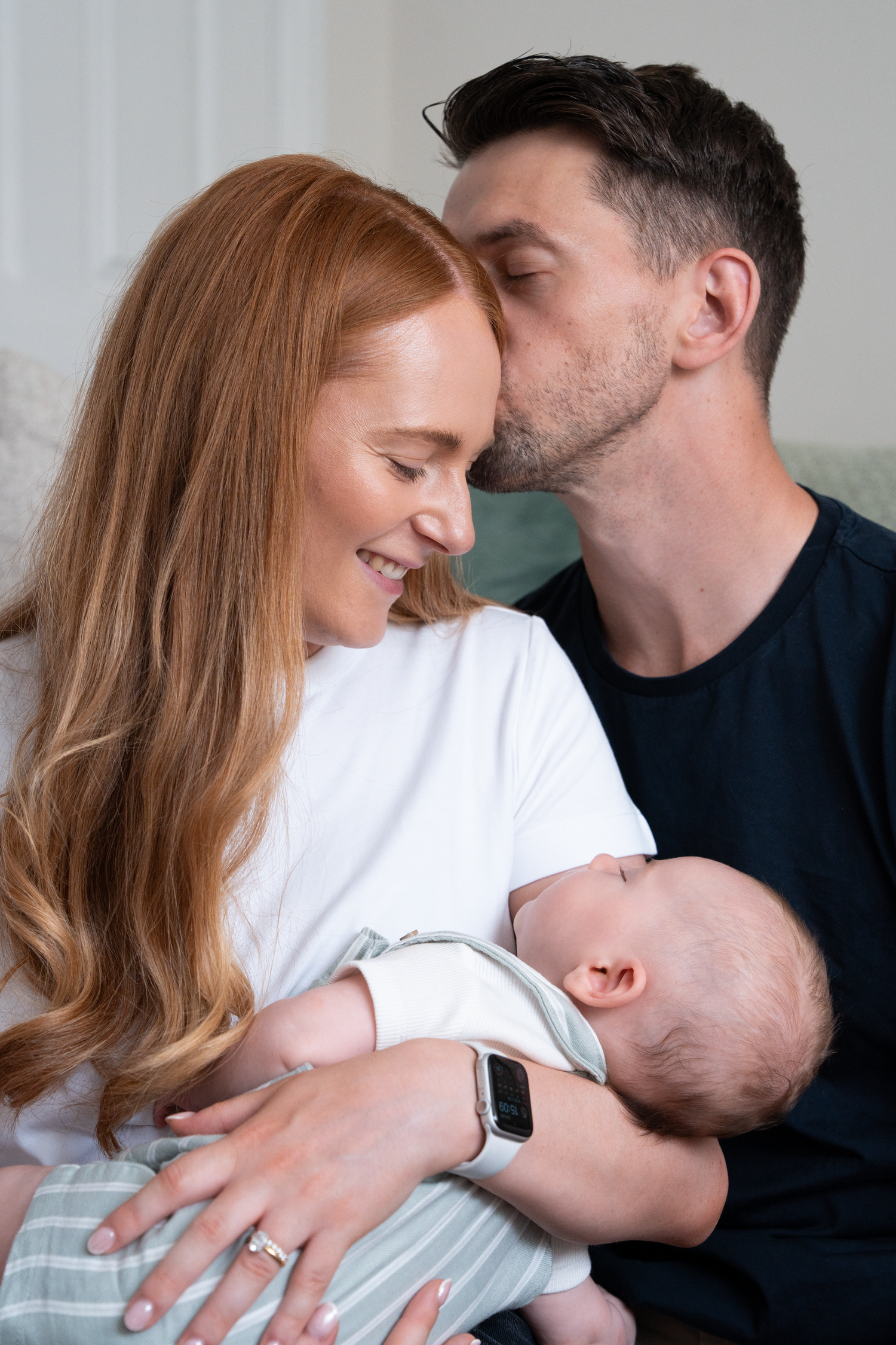 A happy couple shares a tender moment kissing while holding their newborn baby at home.