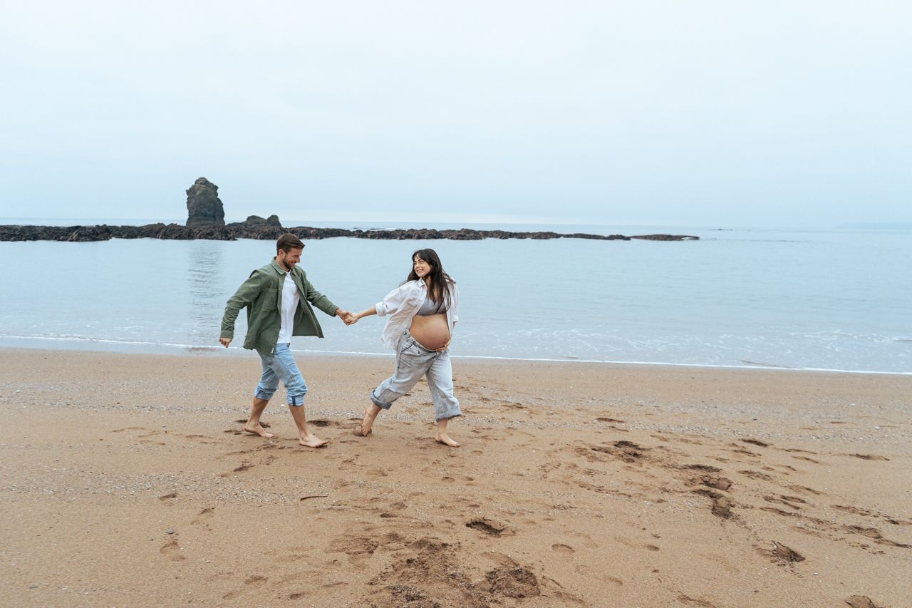 Pregnant couple running and holding hands playfully along sandy beach shoreline.