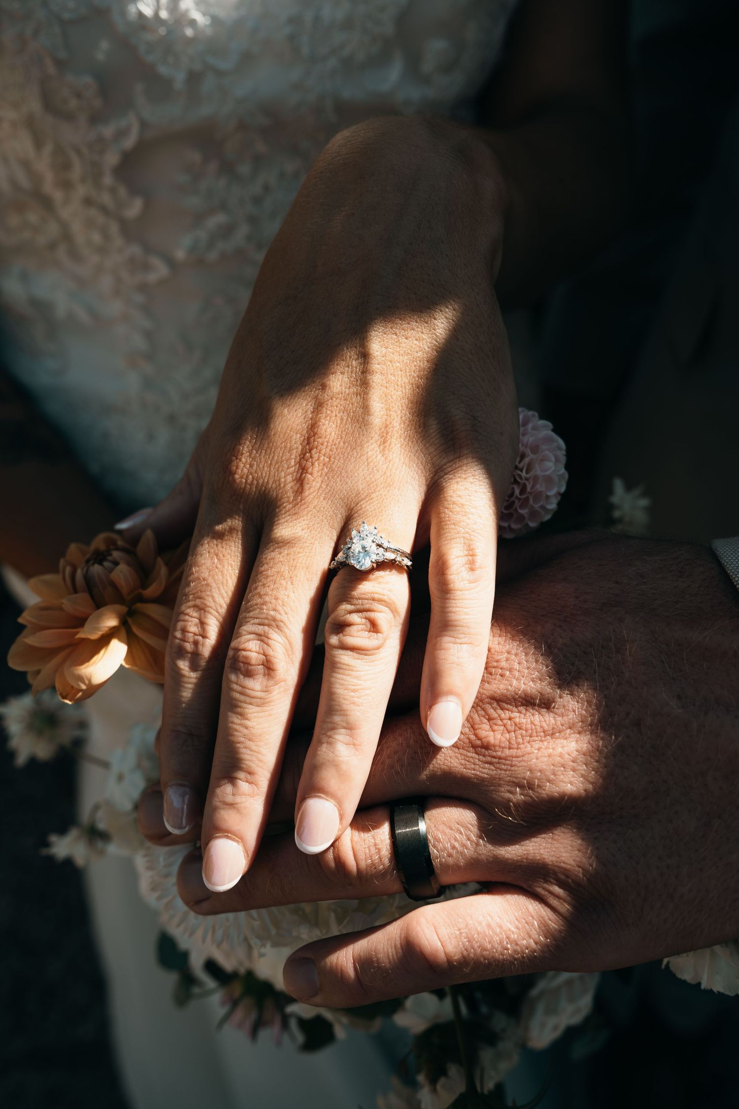 Newlywed couple displaying wedding rings, bride wearing diamond engagement ring, groom with black band, sunlight casting shadows.