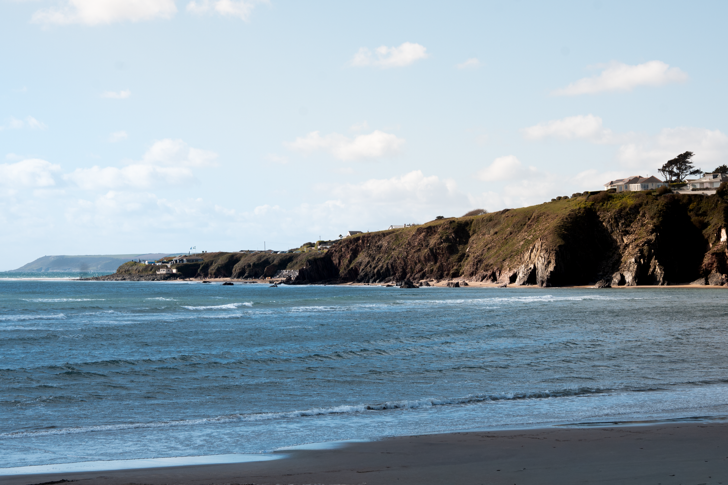 Scenic coastal cliffside view with rocky shoreline, blue ocean waves, and green headland under partly cloudy sky.