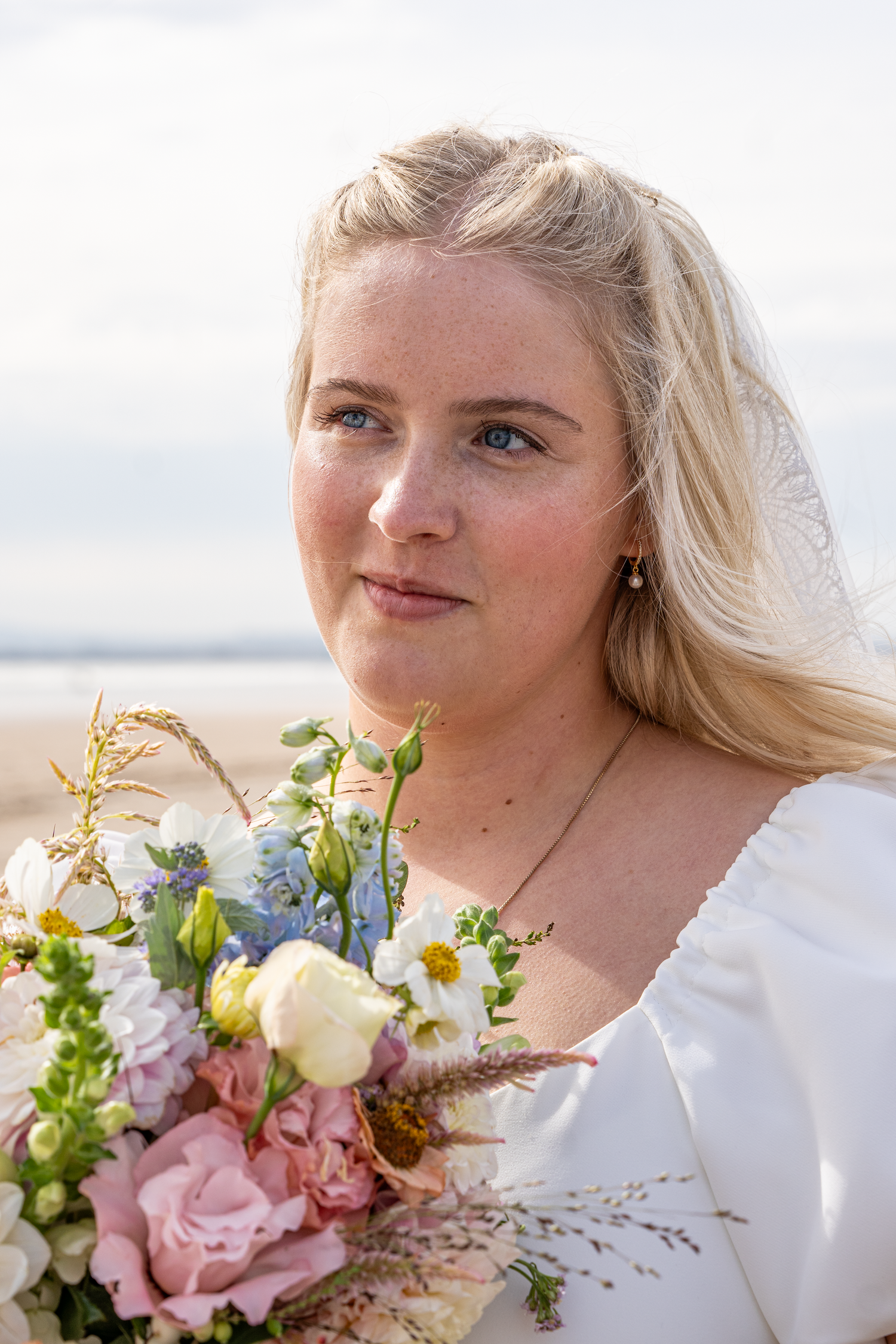 Portrait of blonde bride in white dress holding pastel bouquet of pink roses and blue flowers on beach.
