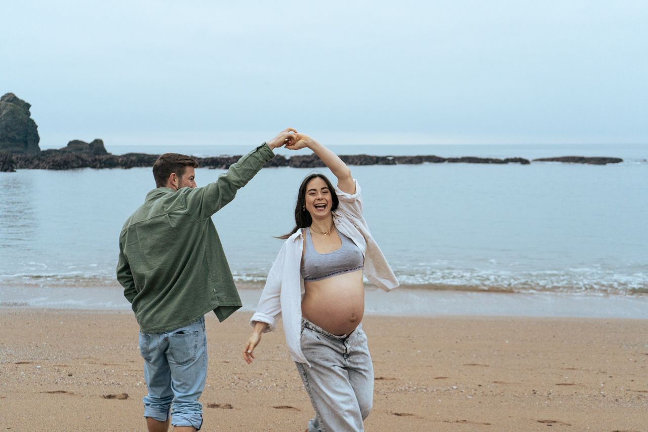 Happy pregnant couple dancing on sandy beach, man spinning woman as ocean waves roll in behind them.