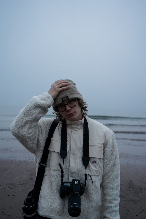 Young photographer with camera at foggy beach wearing white fleece jacket and beige Dickies beanie.
