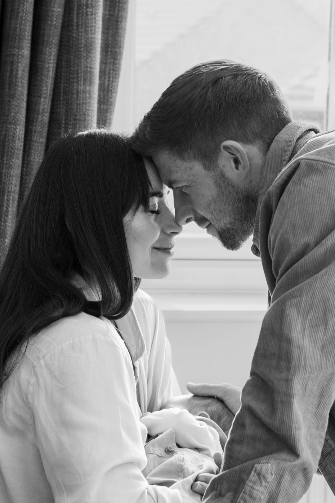 Intimate black and white portrait of couple touching foreheads tenderly by window.
