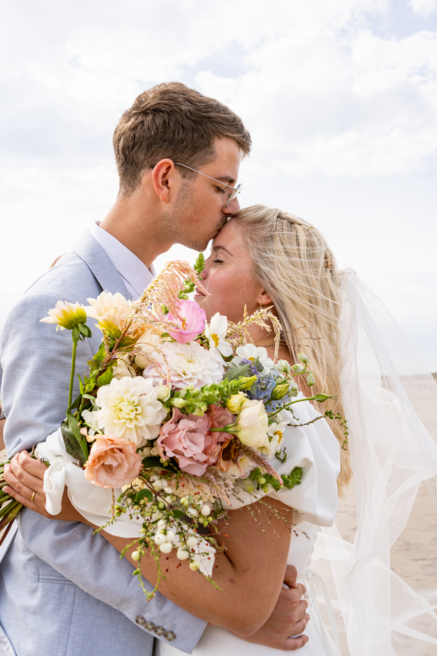 Bride and groom kissing while holding a colorful pastel wedding bouquet on a beach under cloudy skies.