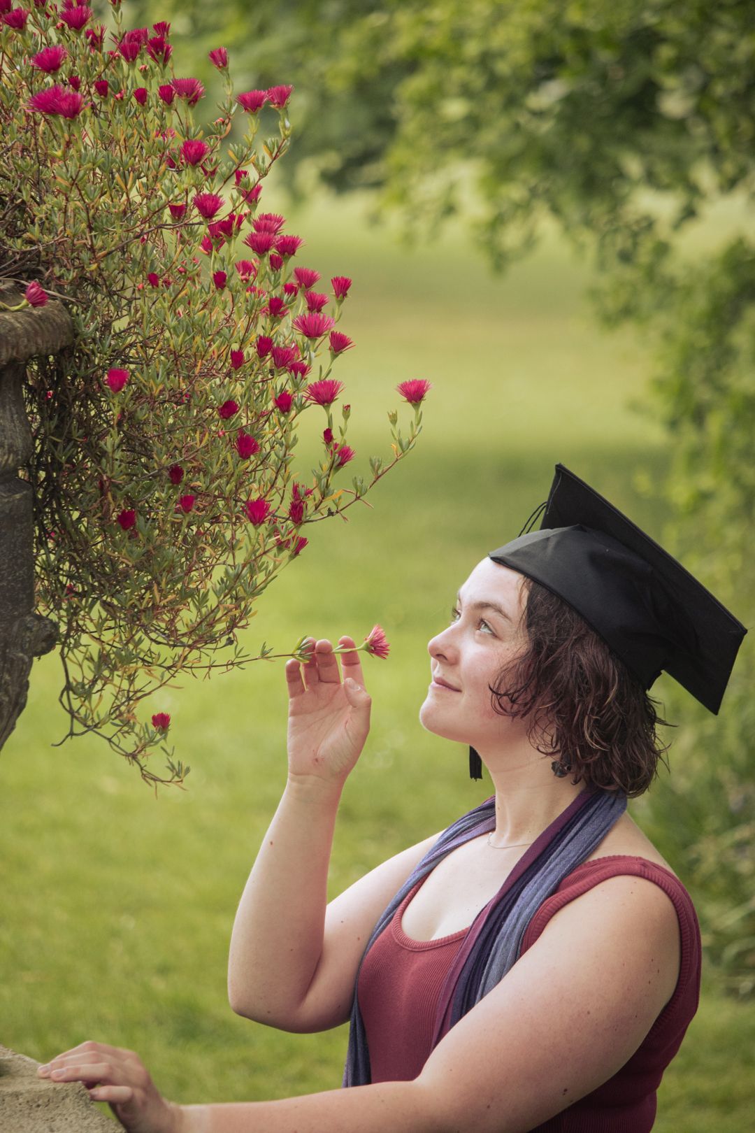 A young woman in a graduation cap admires red flowers on a bush in a green park.