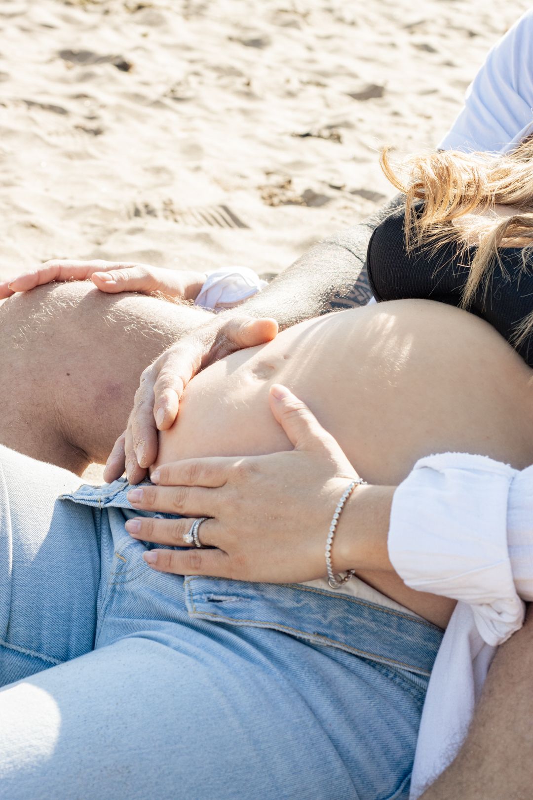 Close-up of couple's hands tenderly holding pregnant belly on sunny beach.