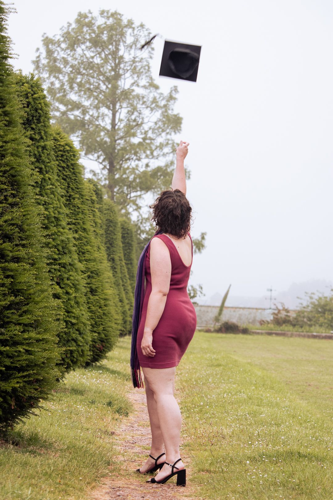 Graduate in burgundy dress joyfully tosses graduation cap into misty sky along garden hedge path.