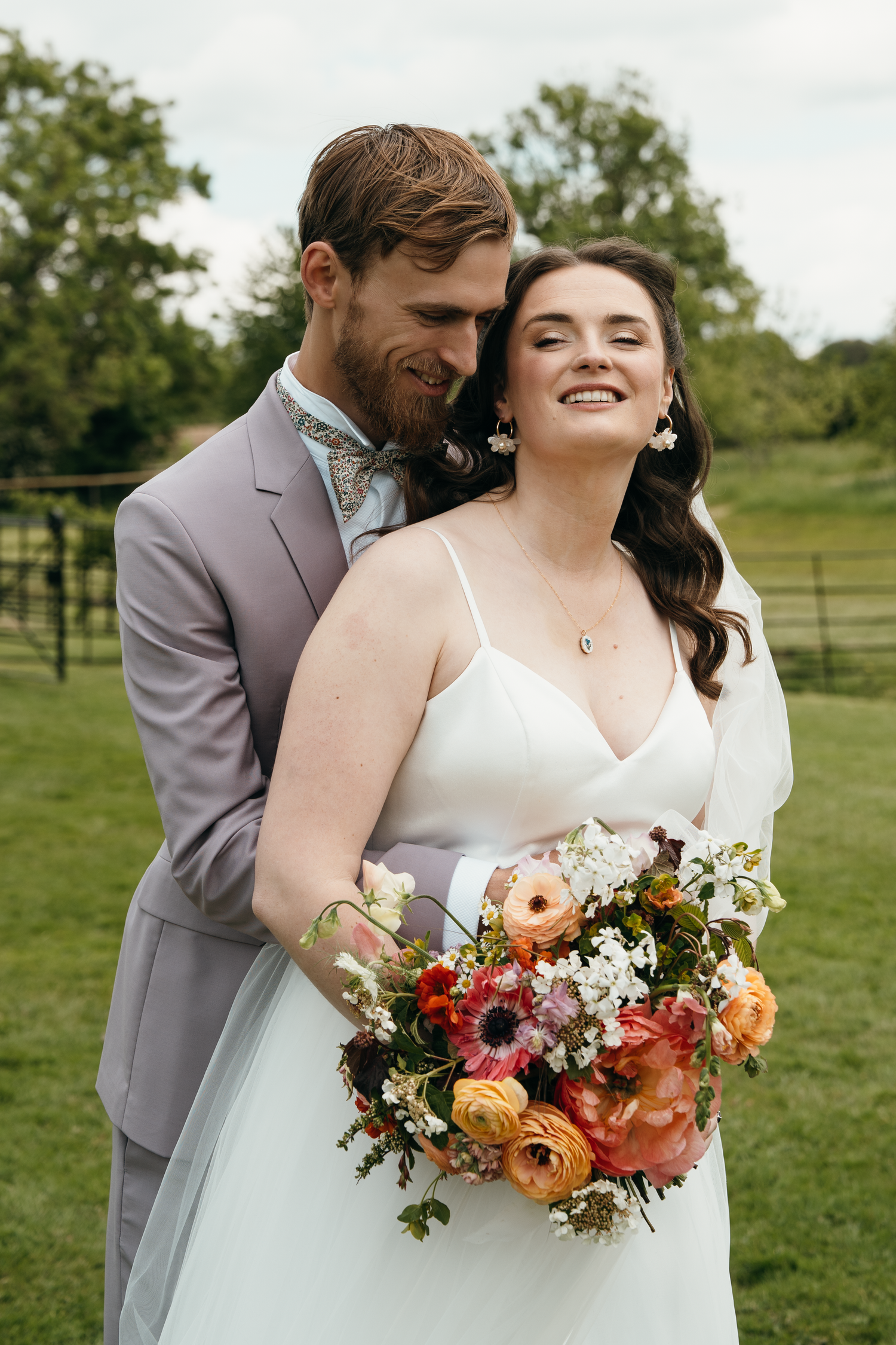 A smiling bride holding a colorful bouquet embraces a groom in a gray suit outdoors at a farm wedding.