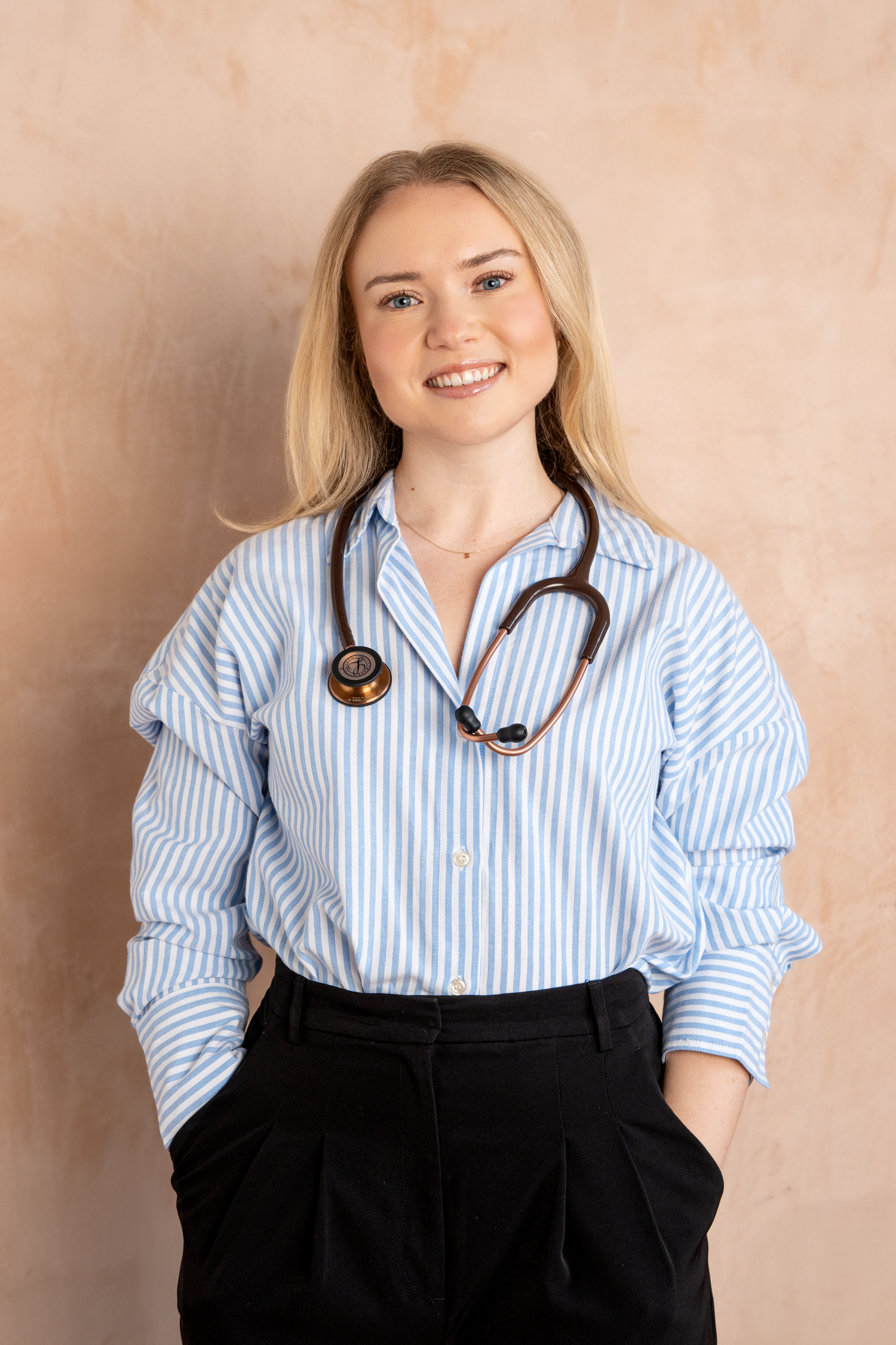 Smiling blonde female doctor wearing a blue striped shirt and stethoscope, standing confidently with hands in pockets.