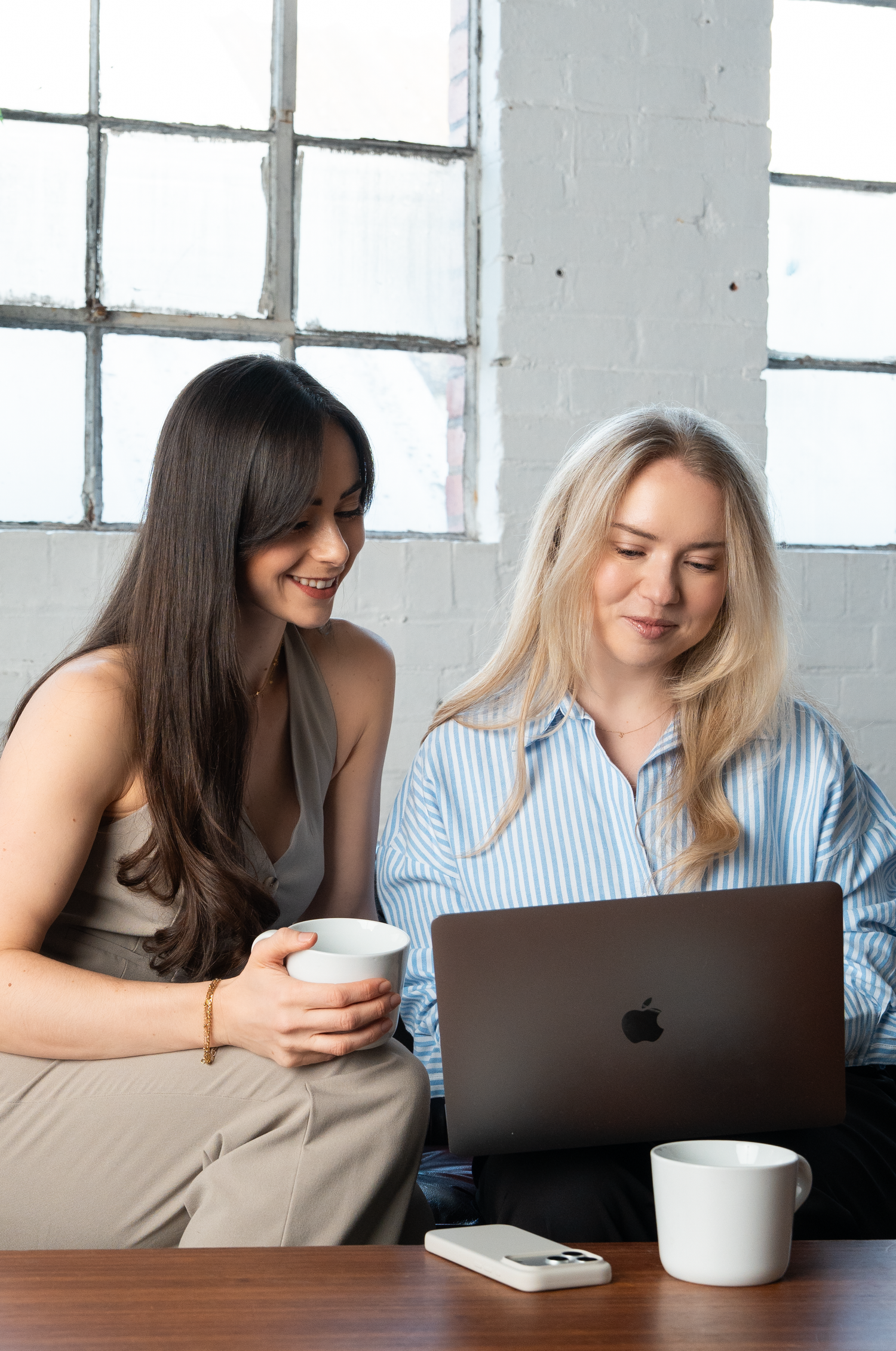 Two women collaborating on a MacBook laptop in a bright industrial office, one holding a coffee cup, smiling together.