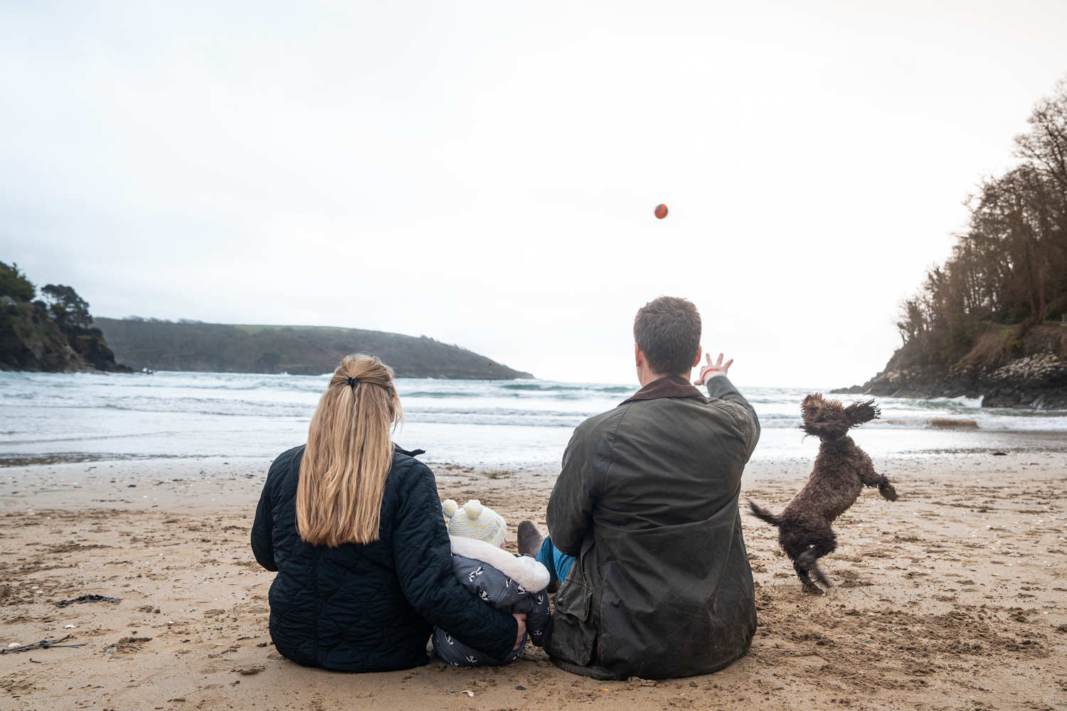 Couple sitting on beach with dog jumping to catch a red ball tossed in the air at sunset.