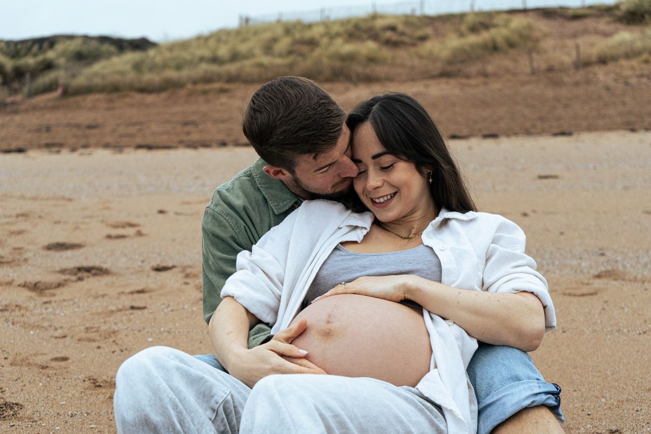 Loving couple sitting on beach sand, man embracing pregnant woman from behind, both smiling warmly.