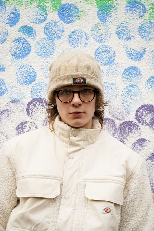 Young person in Dickies beanie and white Dickies jacket posing against colorful dotted wall.