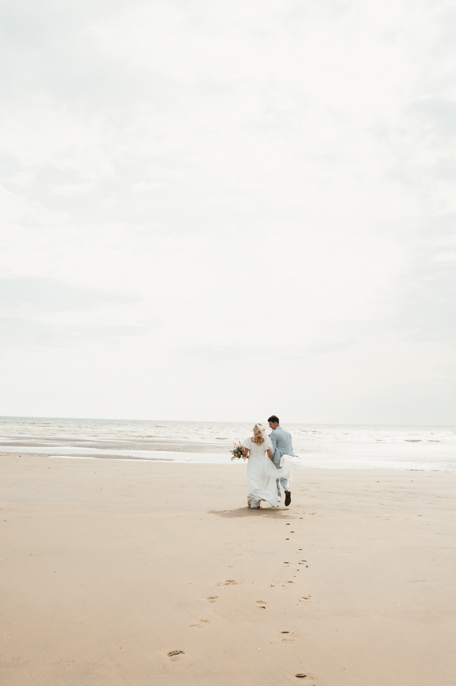 Bride and groom walking hand in hand on a sandy beach, leaving footprints, with ocean waves and overcast sky behind them.