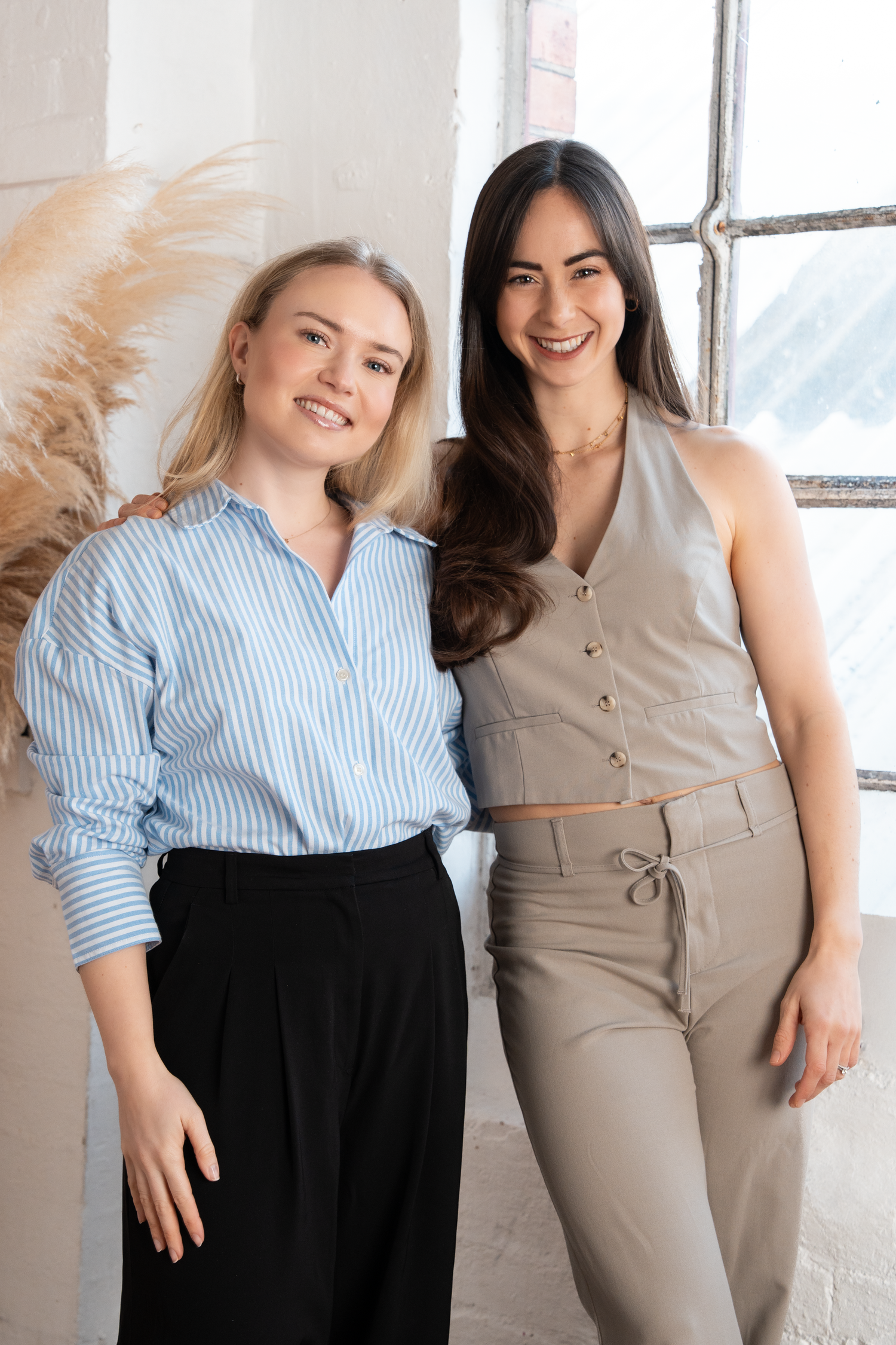 Two smiling women posing together, one in a blue striped shirt and one in a beige halter top and pants.