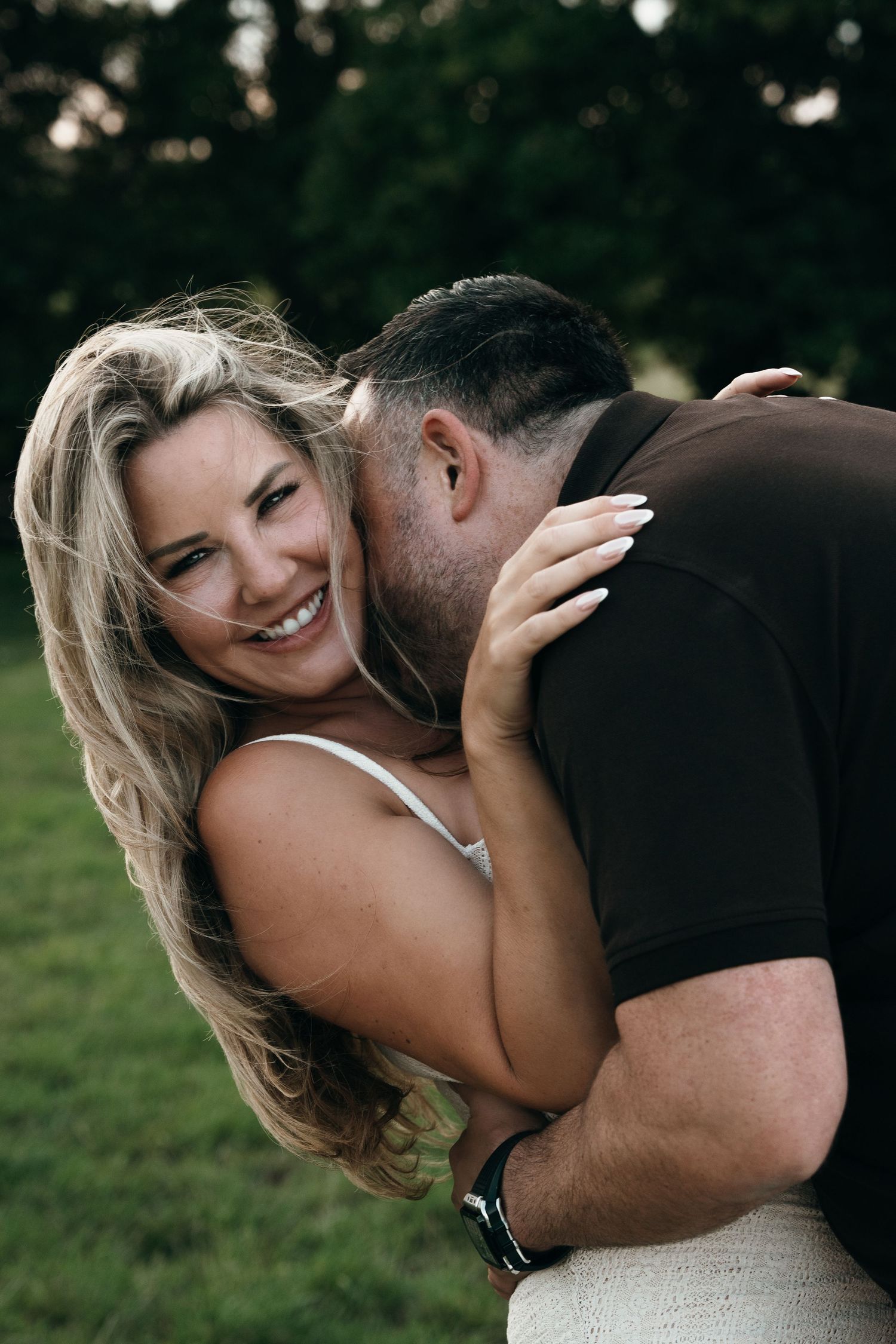 A smiling blonde woman embraces a man kissing her cheek outdoors in a lush green park setting.