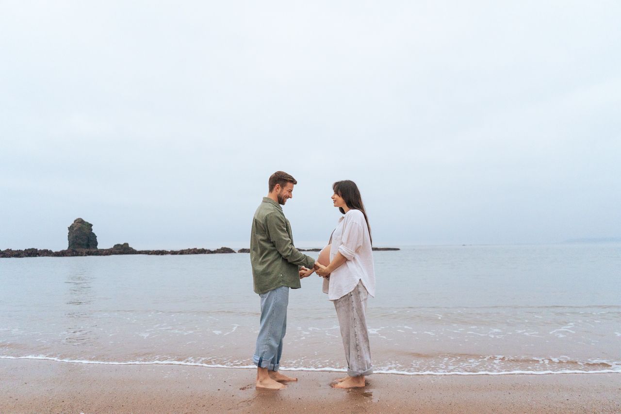 Couple holding hands facing each other on calm beach with overcast sky.