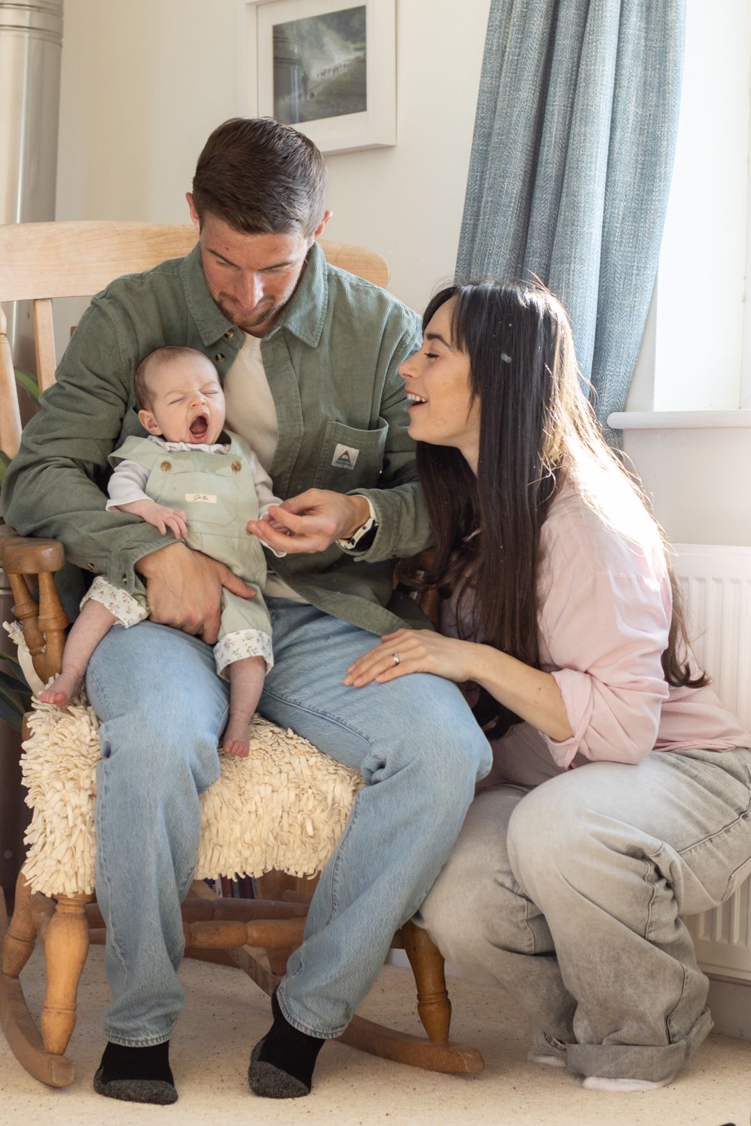 Young couple laughing together with yawning baby in wooden rocking chair at home.