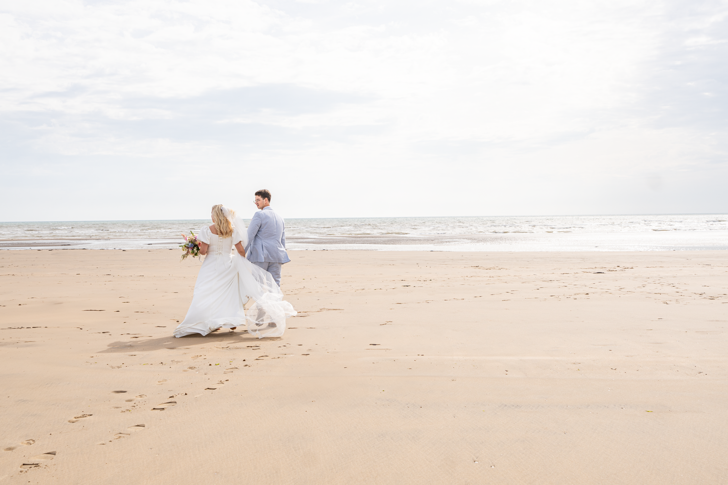 Bride and groom walking hand in hand along a wide sandy beach leaving footprints in the sand.