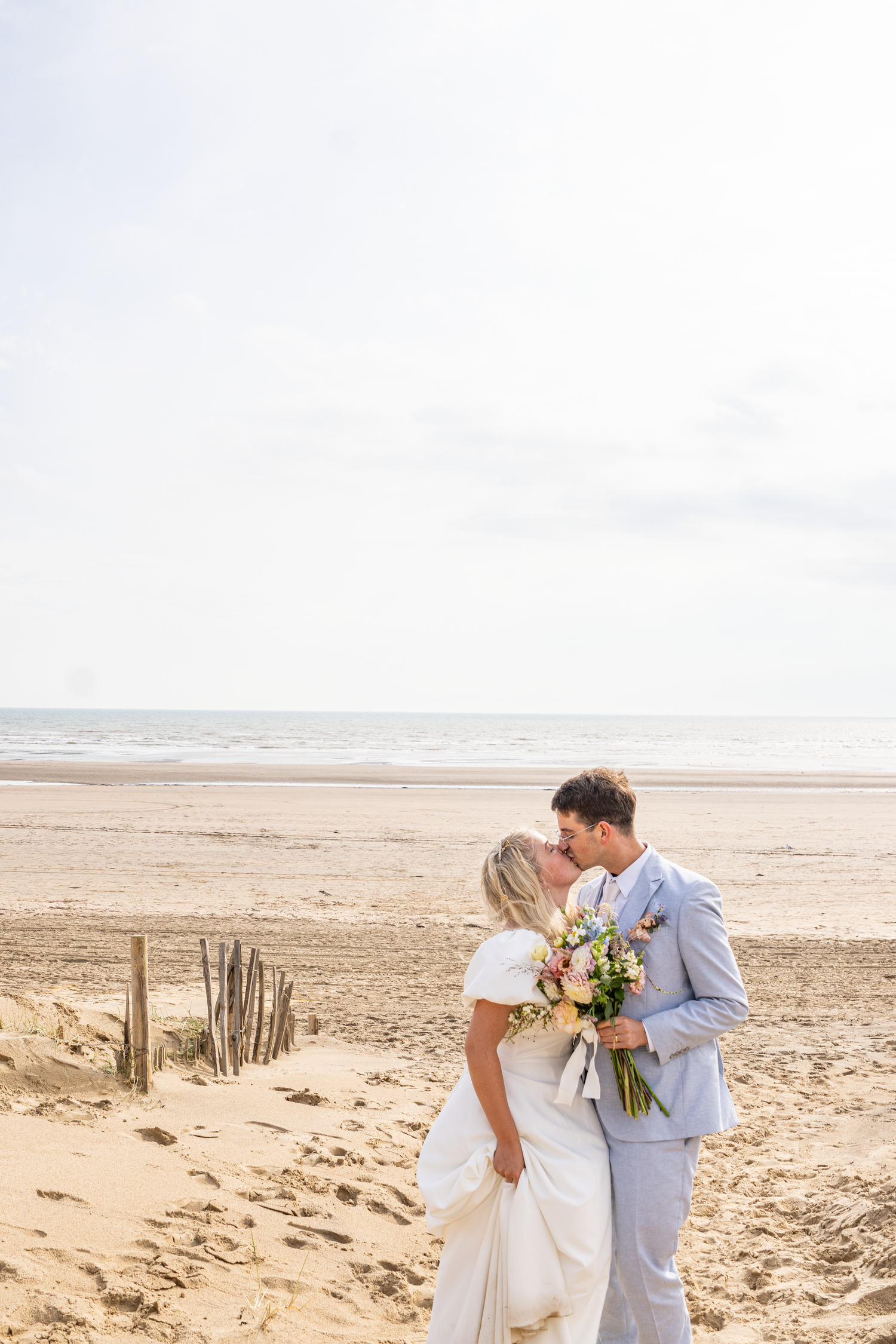 Bride and groom kissing on a sandy beach holding a colorful bouquet, with the ocean in the background.