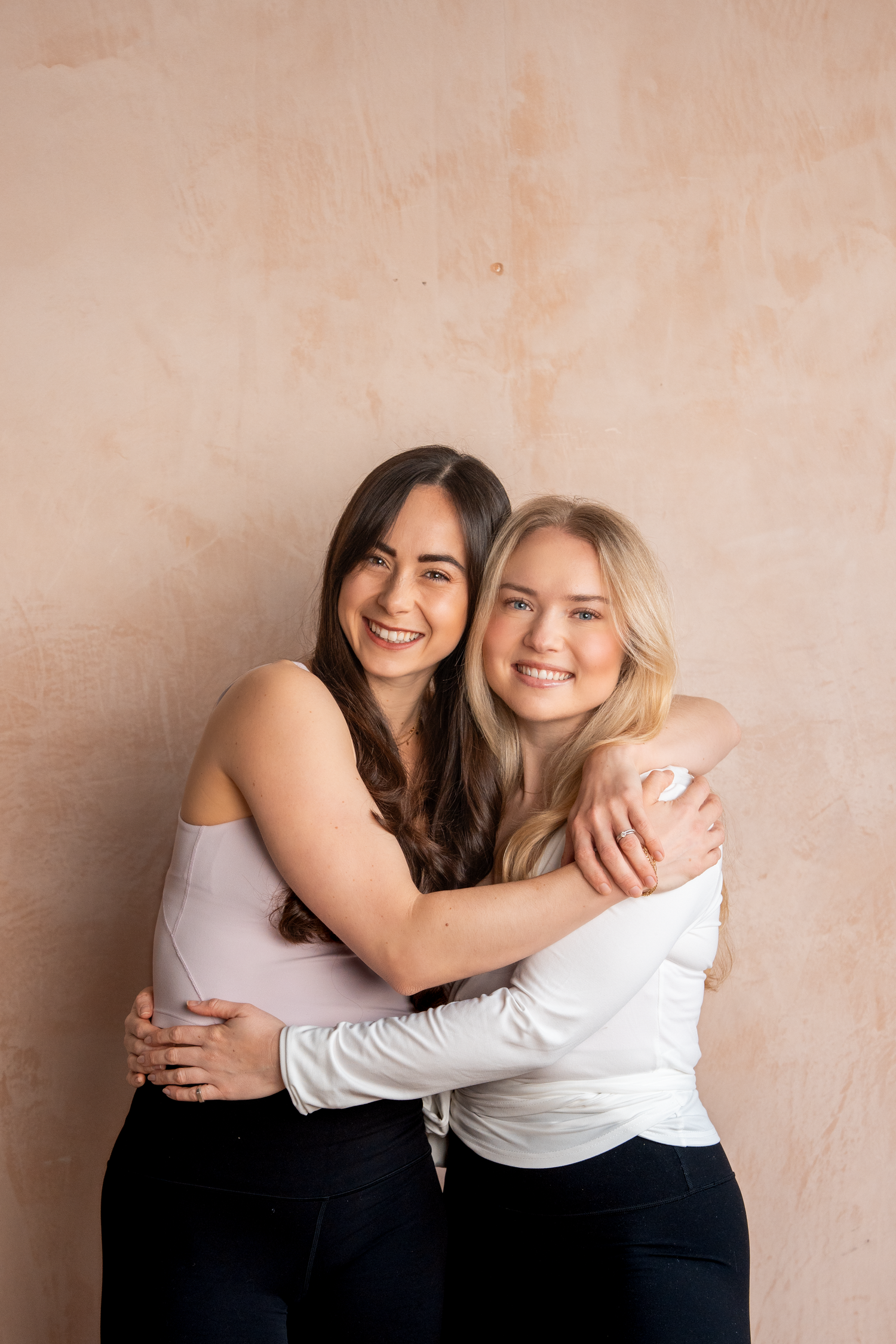 Two smiling women embracing warmly in casual clothing against a neutral beige background.