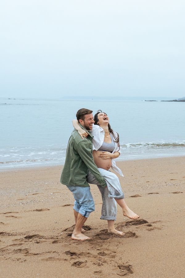 Joyful pregnant couple laughing and embracing on a sandy beach with calm ocean behind them.
