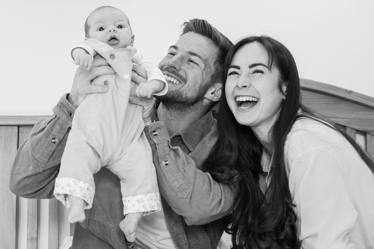 Black and white photo of laughing parents lifting happy baby on wooden bench.