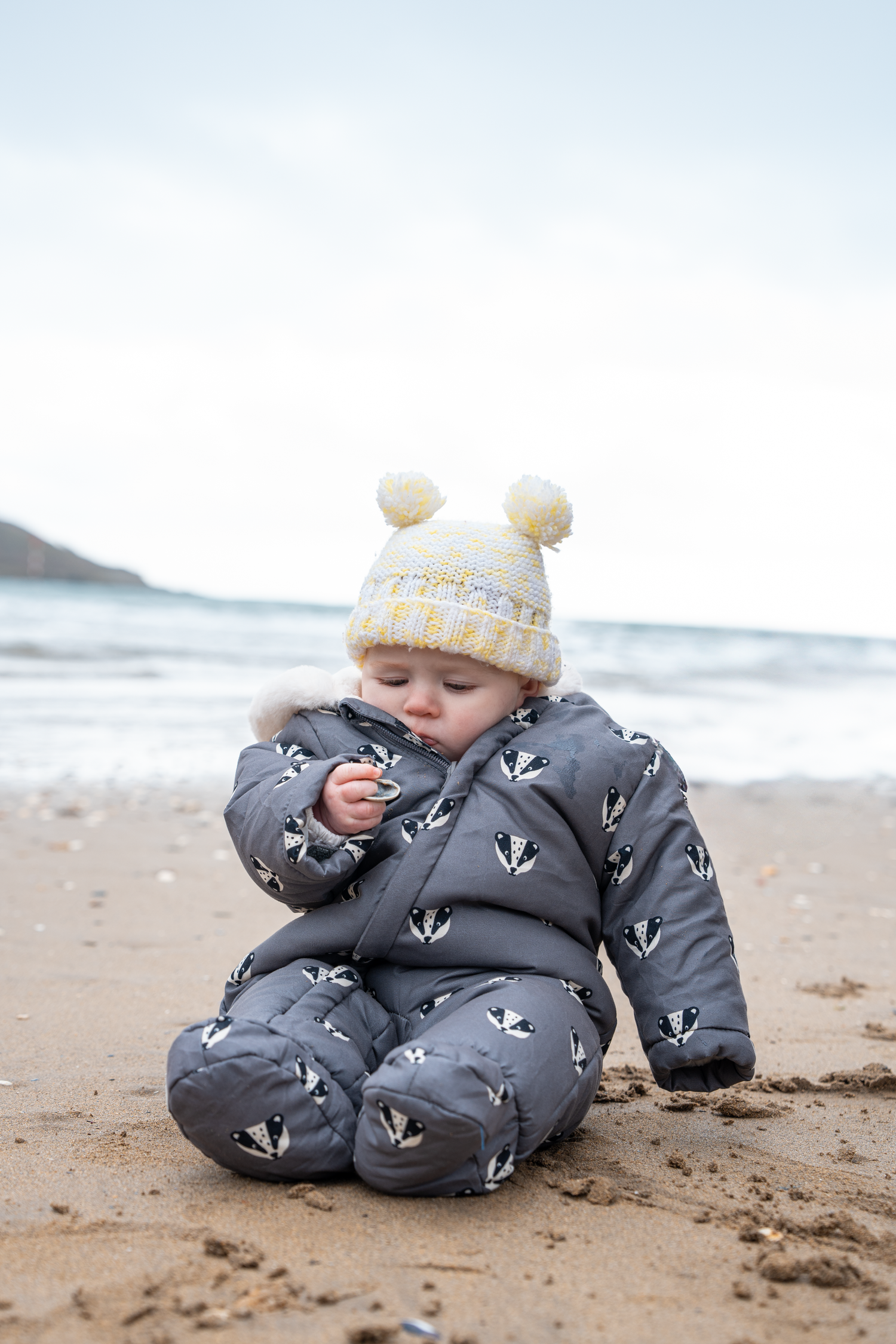 Baby in grey badger-print snowsuit and yellow pompom hat sitting on sandy beach exploring surroundings.
