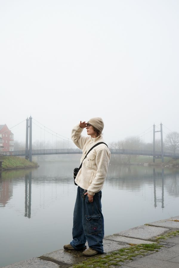 Person in white fleece jacket and beanie shielding eyes by misty river with suspension bridge.