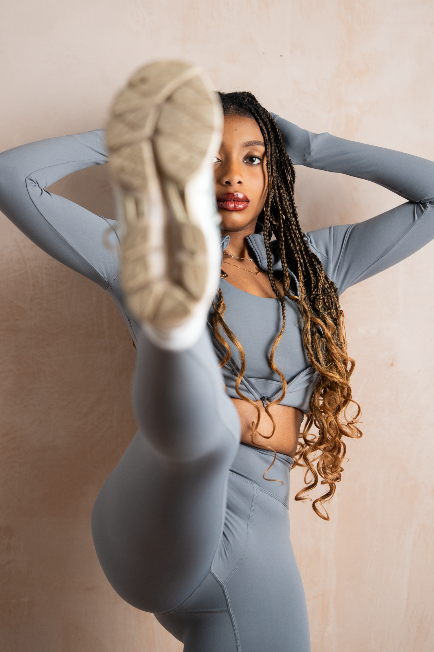 A woman in a grey athletic set performs a high kick pose against a lime wash wall, showcasing flexibility and sportswear.