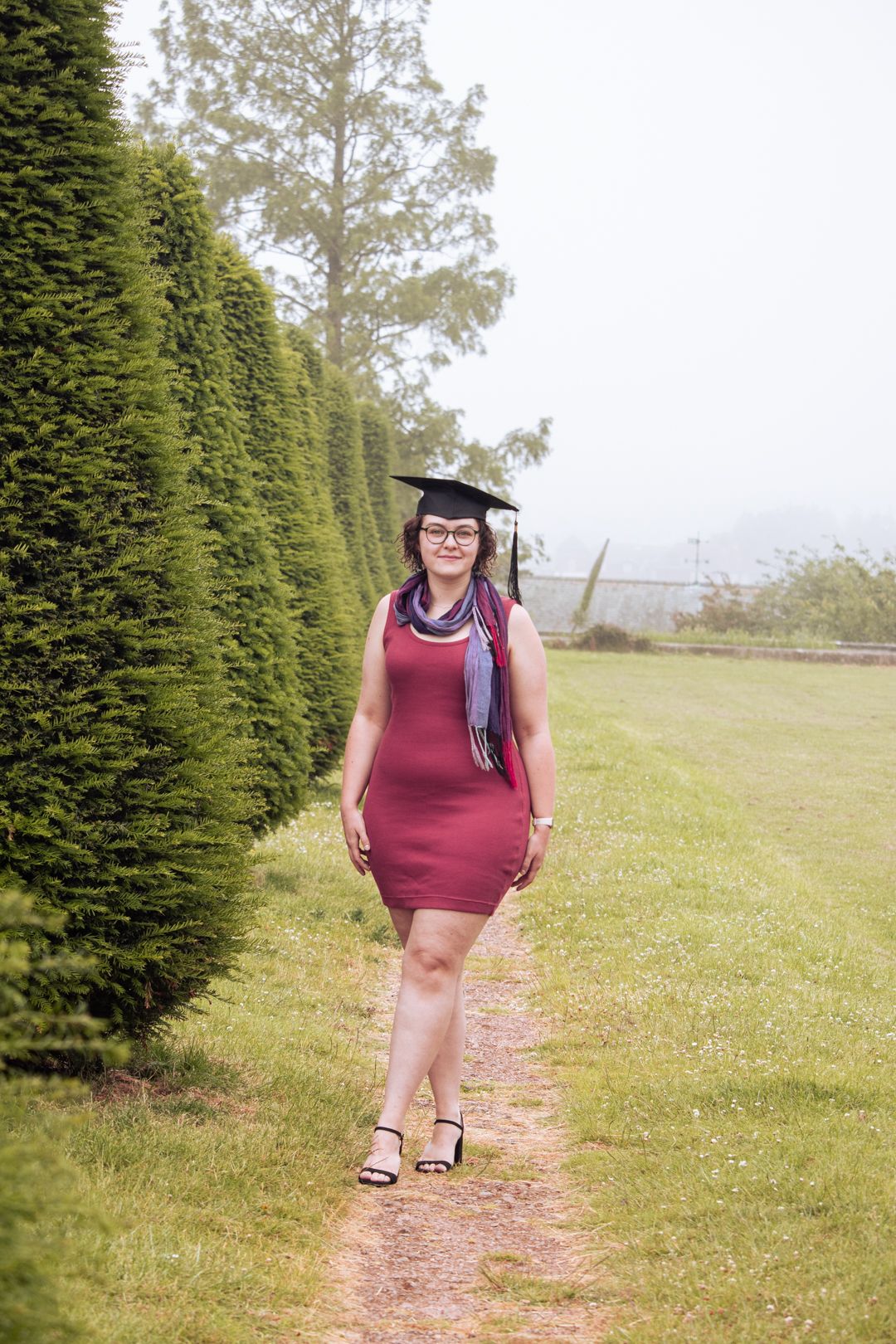 Graduate in burgundy dress and graduation cap walks along garden path flanked by tall green hedges.