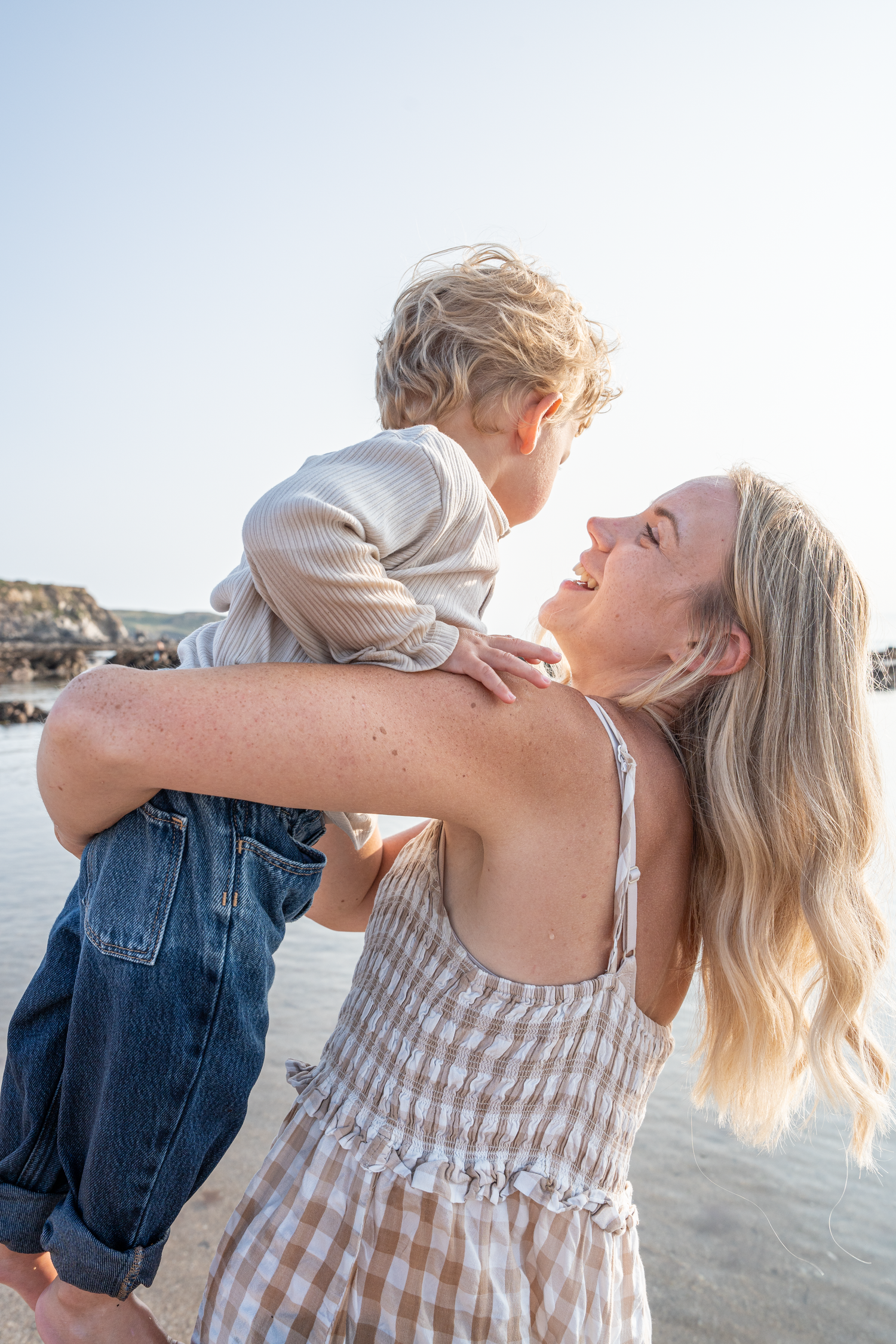 Joyful blonde mother lifting and embracing her toddler son on a sunny beach.