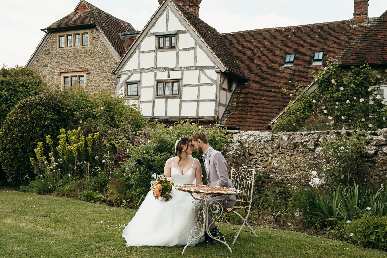 Bride and groom sit at ornate garden table outside historic Tudor manor house surrounded by lush English cottage garden.