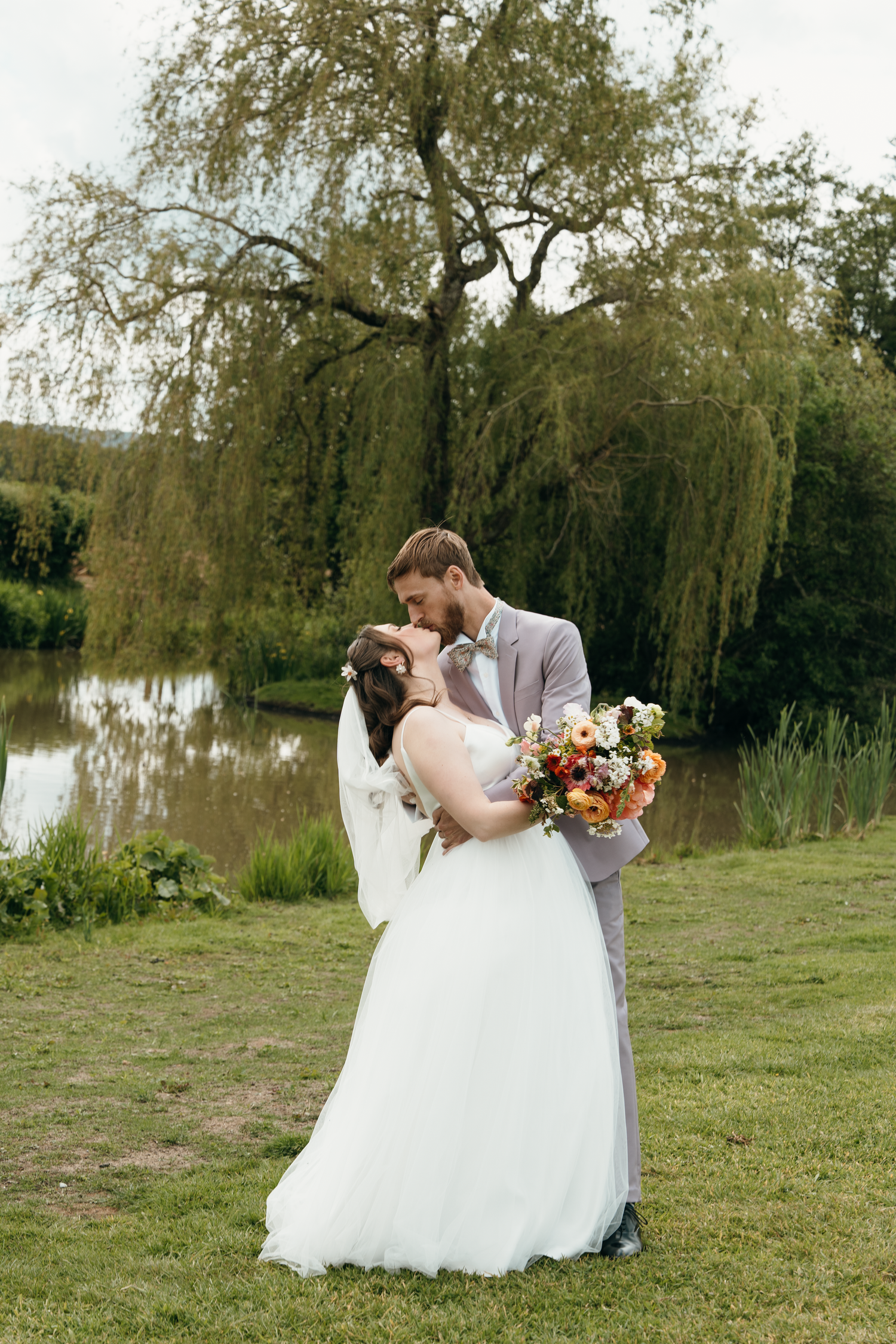 A bride and groom share a romantic kiss by a serene pond, surrounded by lush greenery, as she holds a colorful bouquet.