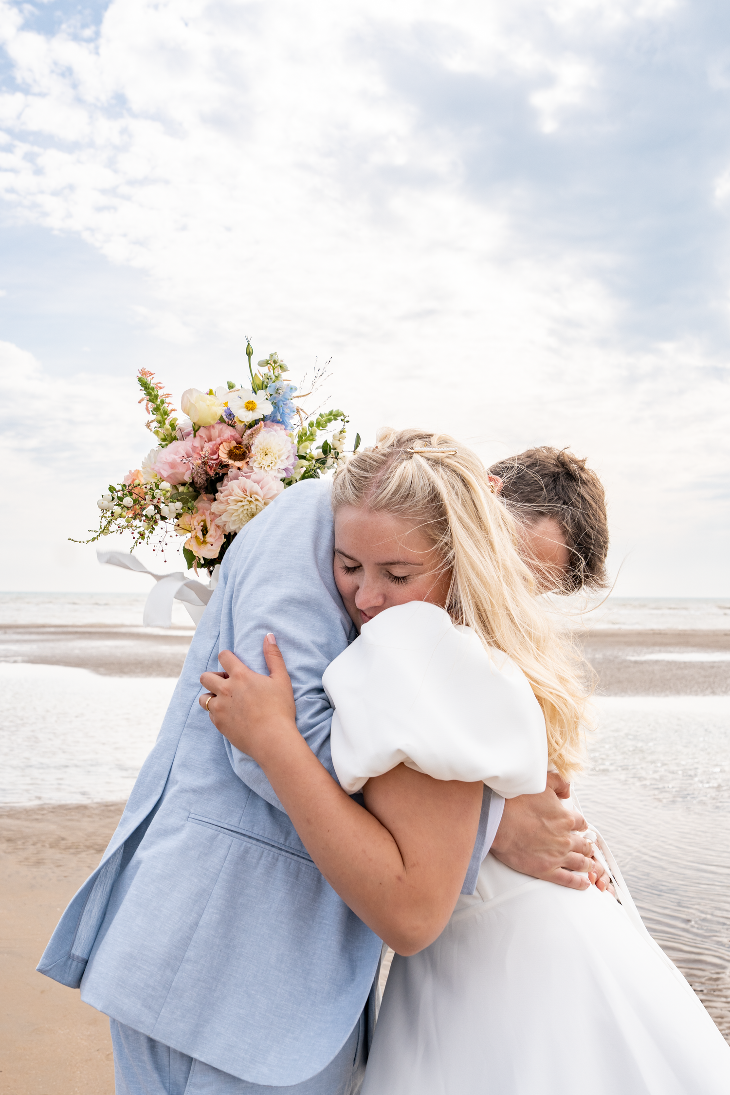 Bride and groom embracing on a beach, bride holding a pastel bouquet, wearing white dress and light blue suit.