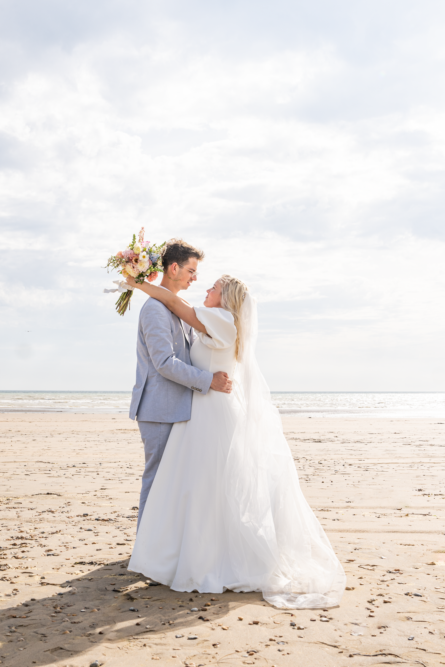 Bride and groom embrace on sandy beach holding colorful bouquet with cloudy sky backdrop on wedding day.