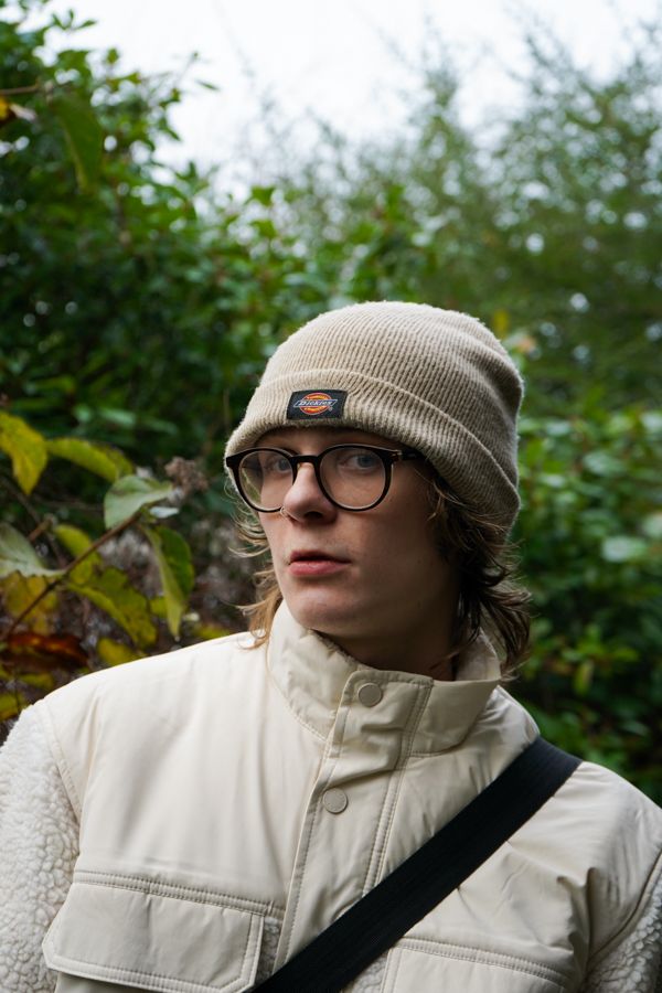Young person in Dickies beanie and white jacket standing before lush green foliage outdoors.