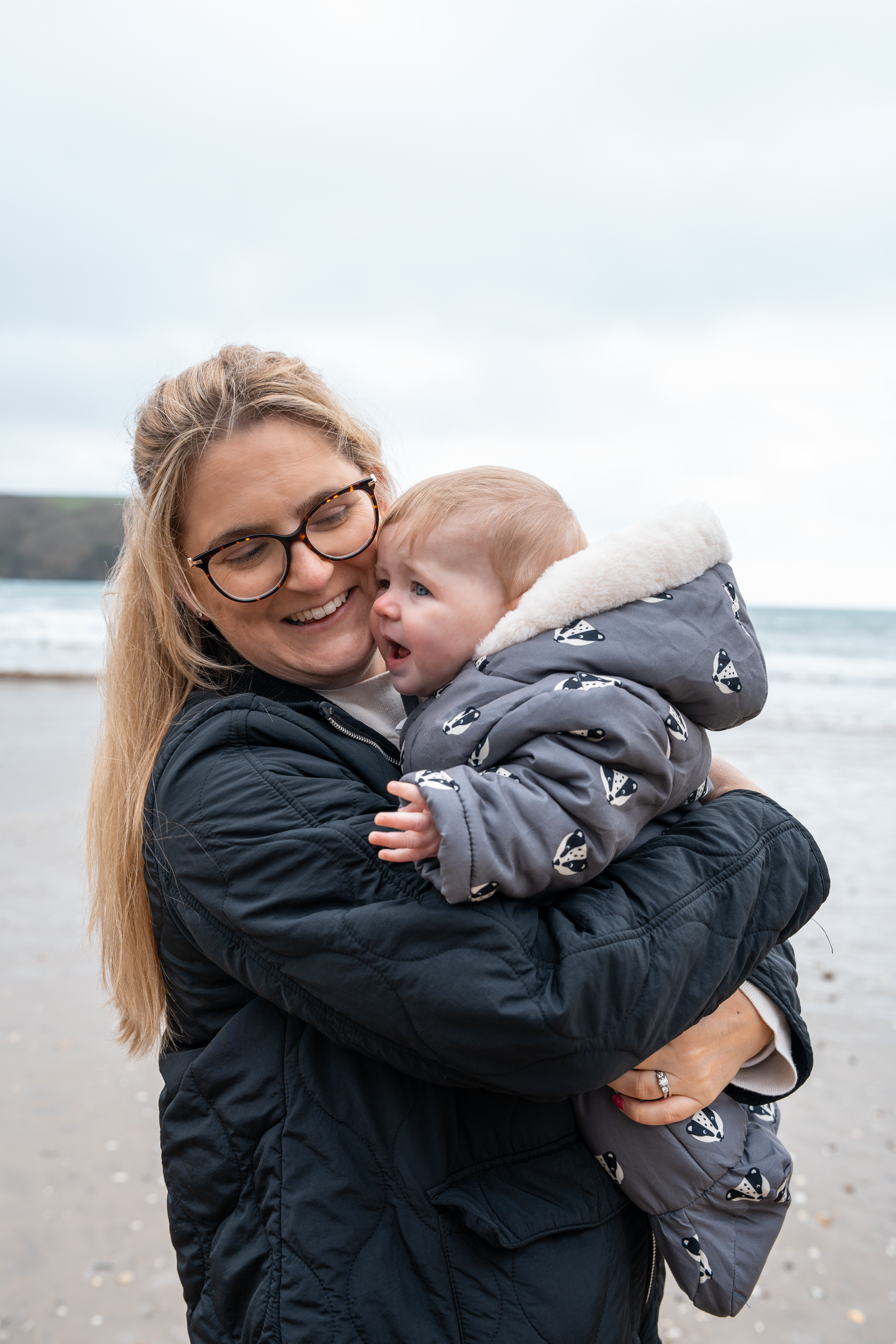 Smiling woman with glasses hugging a baby in a grey patterned snowsuit on a cloudy beach.