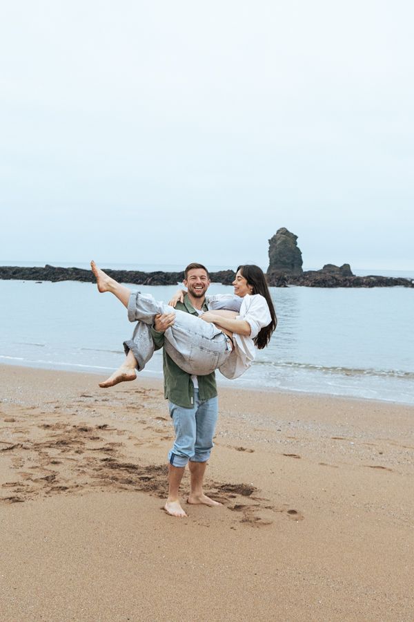 Smiling man carrying pregnant woman in his arms on a sandy beach with rocky ocean backdrop.