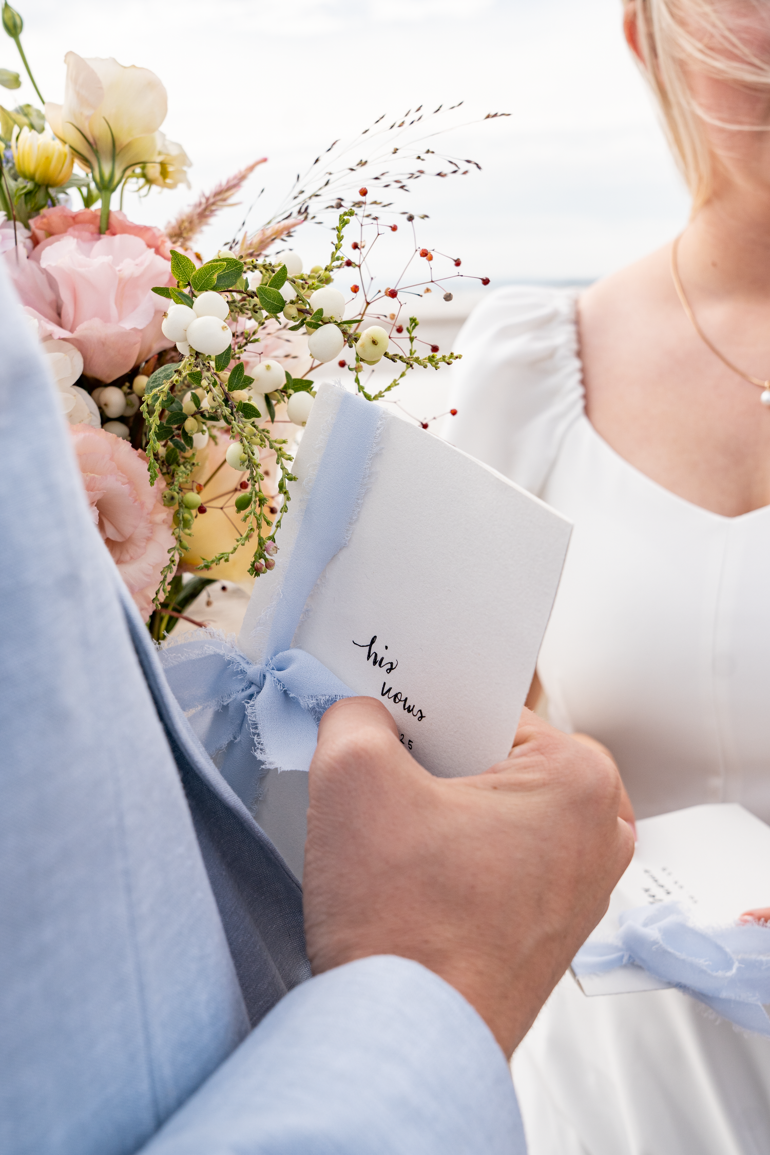 Bride holding wedding vows booklet and bouquet of pink and white flowers beside groom in blue suit.