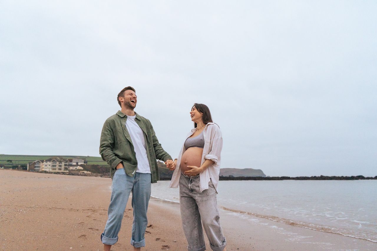 Expecting couple holding hands and smiling on a sandy beach under a cloudy sky.