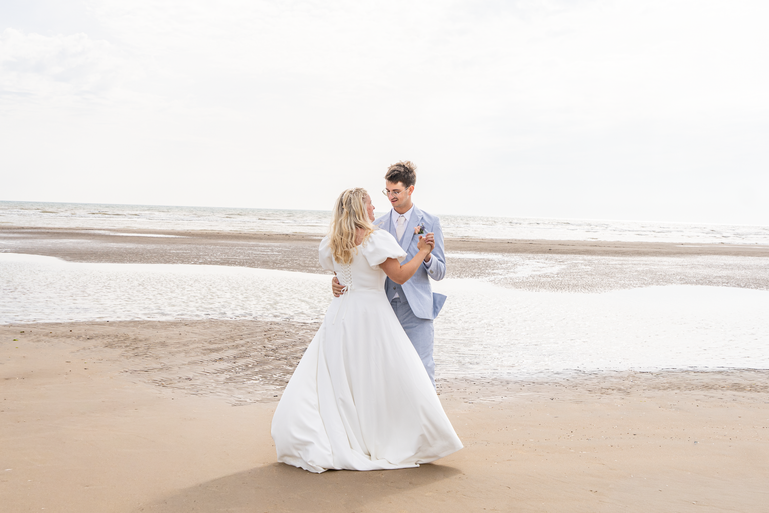 A bride in a flowing white dress and groom dancing romantically on a sandy beach by the ocean.