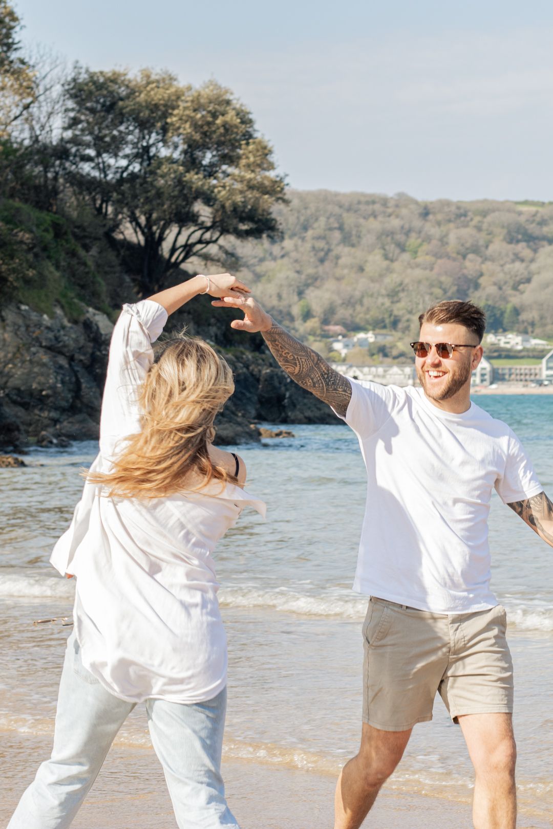 Smiling tattooed man spinning pregnant partner on sunny beach with coastal scenery.