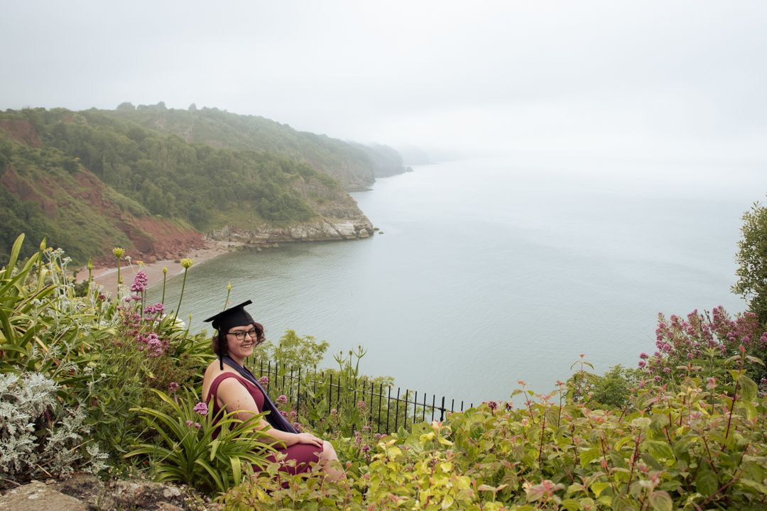 Graduate in graduation cap sits on coastal hillside surrounded by wildflowers with scenic bay view.