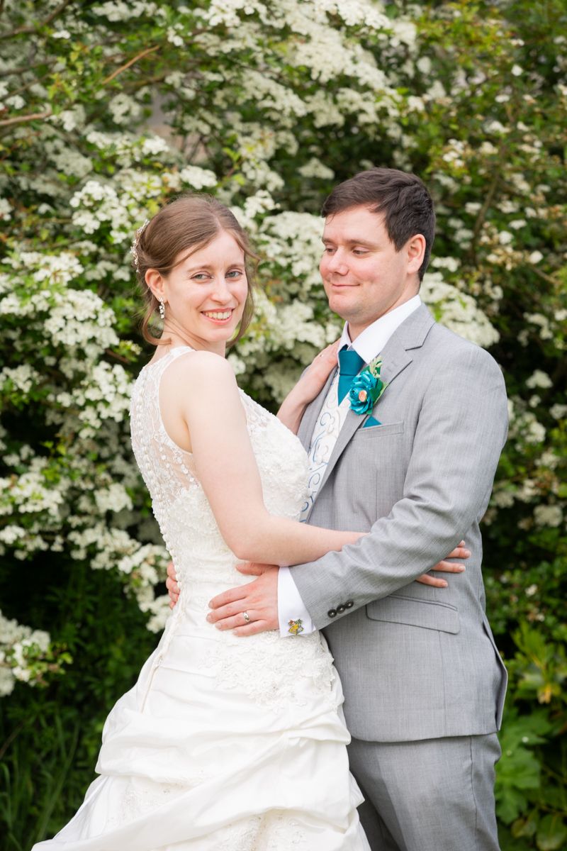 Bride smiling over shoulder while groom embraces her in front of white flowering garden bushes.