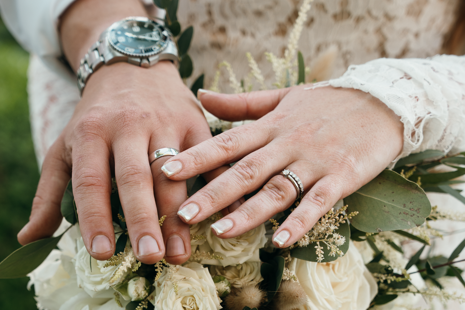 Bride and groom displaying wedding rings on hands resting on a white floral bouquet with eucalyptus greenery.