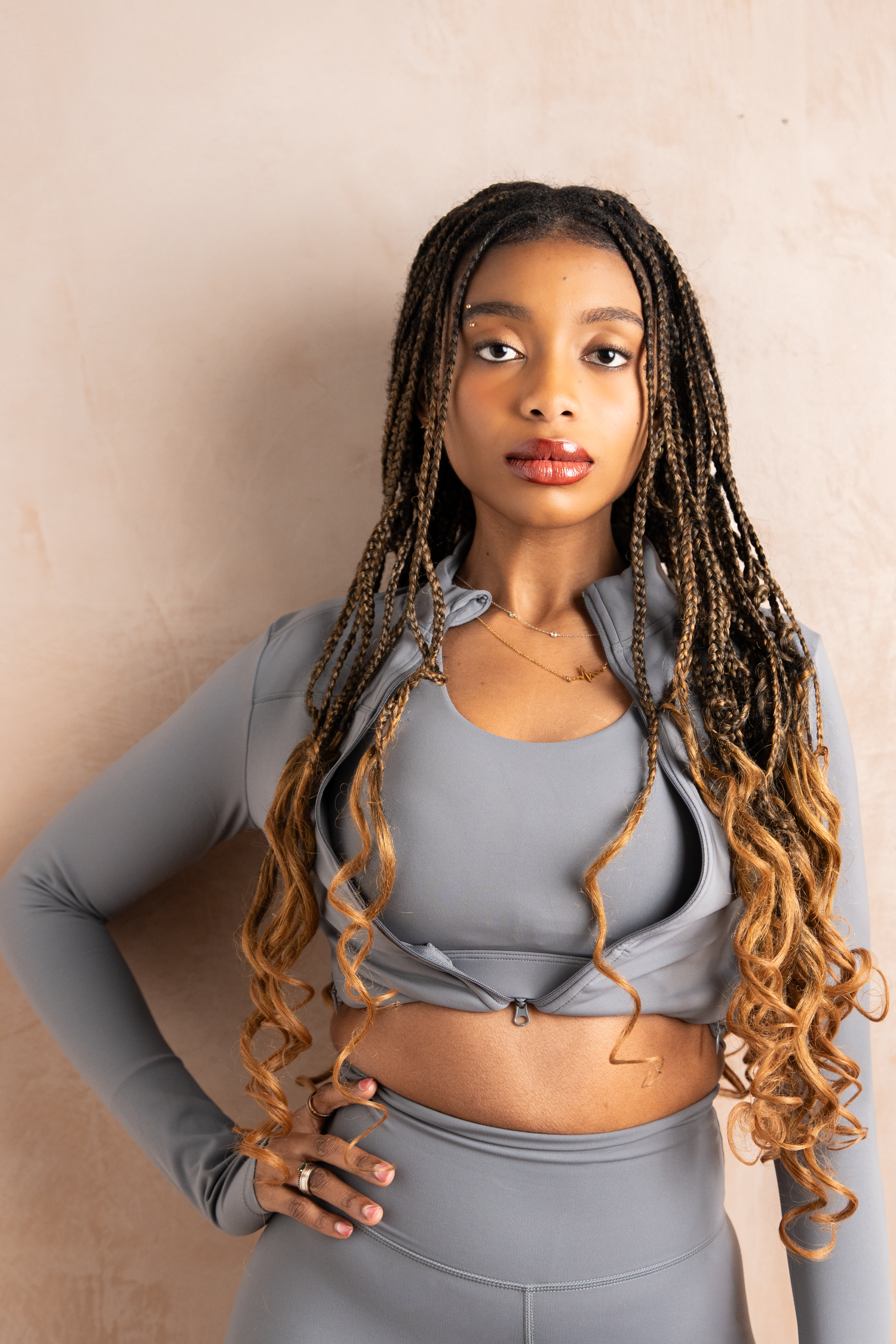 Young woman with long braided hair wearing a grey sports crop top and matching pants, posing confidently against a lime wash wall.